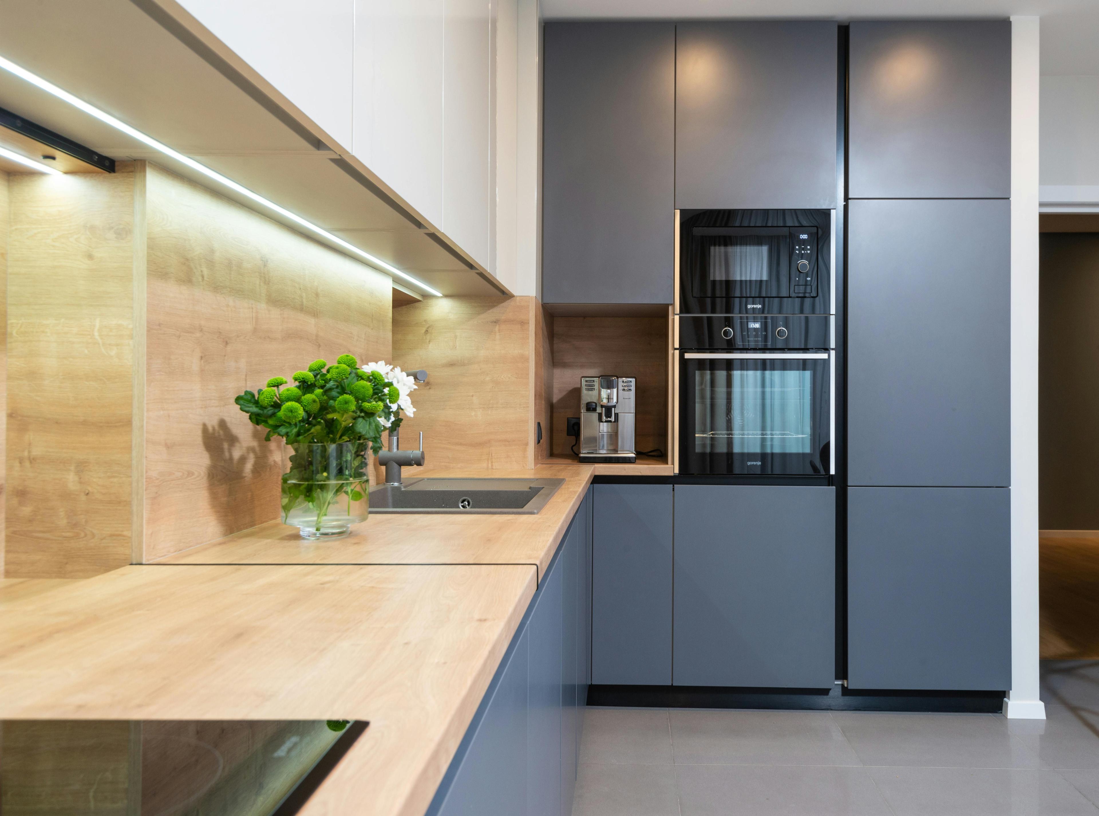 Moderne L-Küche mit grauen Fronten, hellen Holzarbeitsplatten und Rückwand in Holzoptik, integrierter Beleuchtung unter den Hängeschränken sowie schwarzen Einbaugeräten.